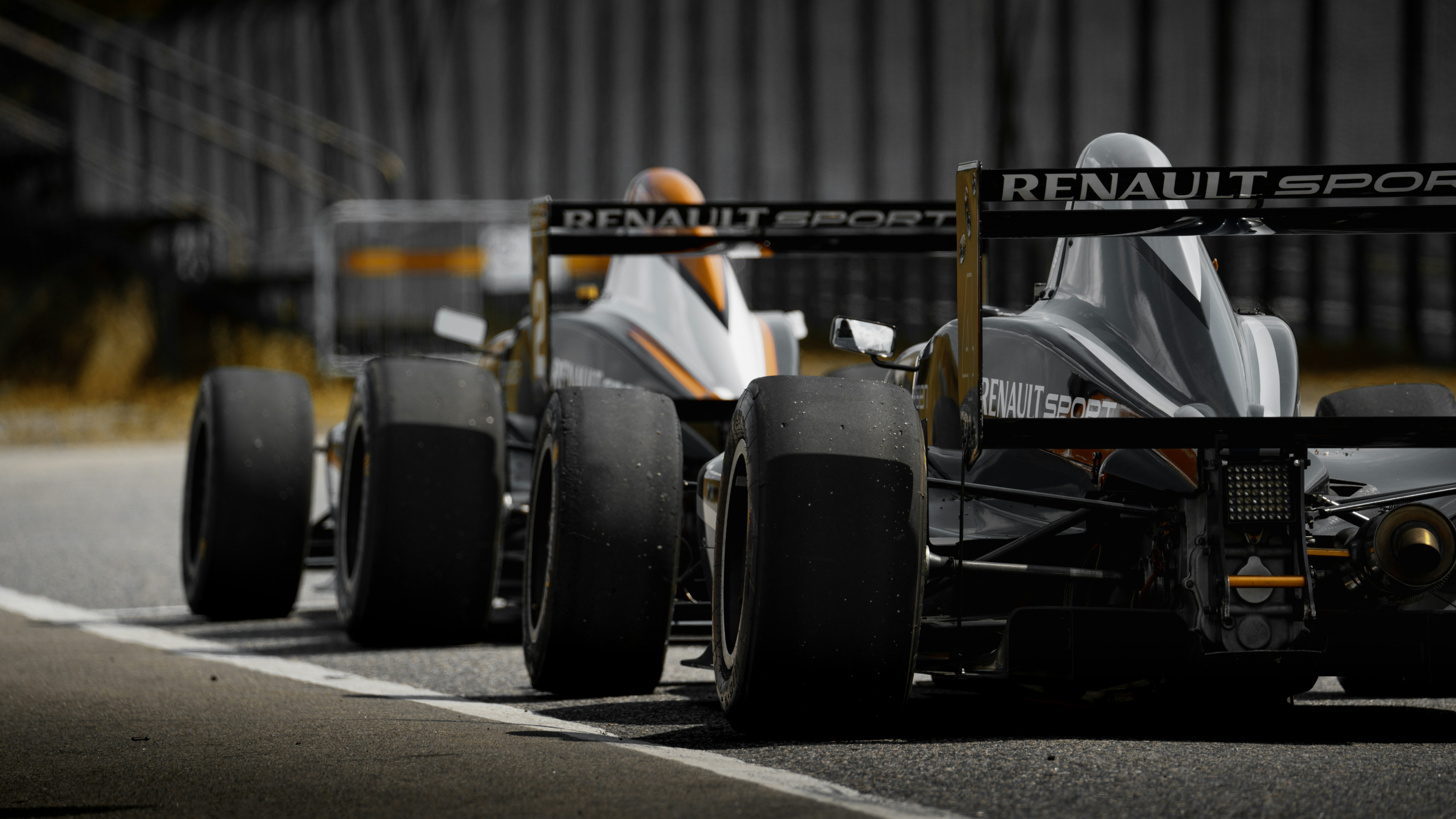Two open-wheel racing cars lined up on a racetrack, viewed from the rear, with large rear tyres, aerodynamic bodywork, and rear wings displaying “Renault Sport”.