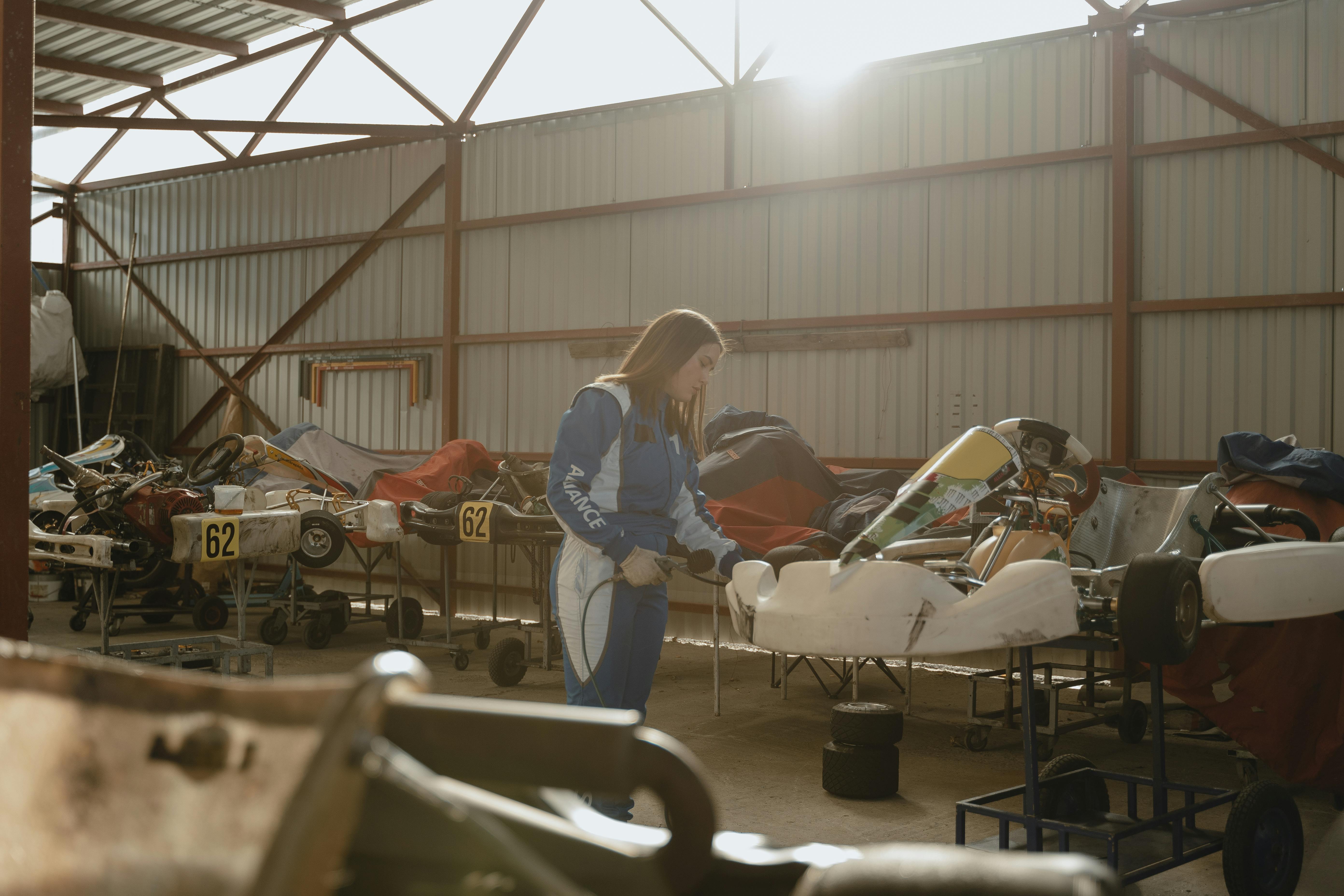 Person in a blue racing suit working on a go-kart inside a motorsport workshop, surrounded by kart chassis, tools, spare parts, and equipment, with sunlight streaming through the building.