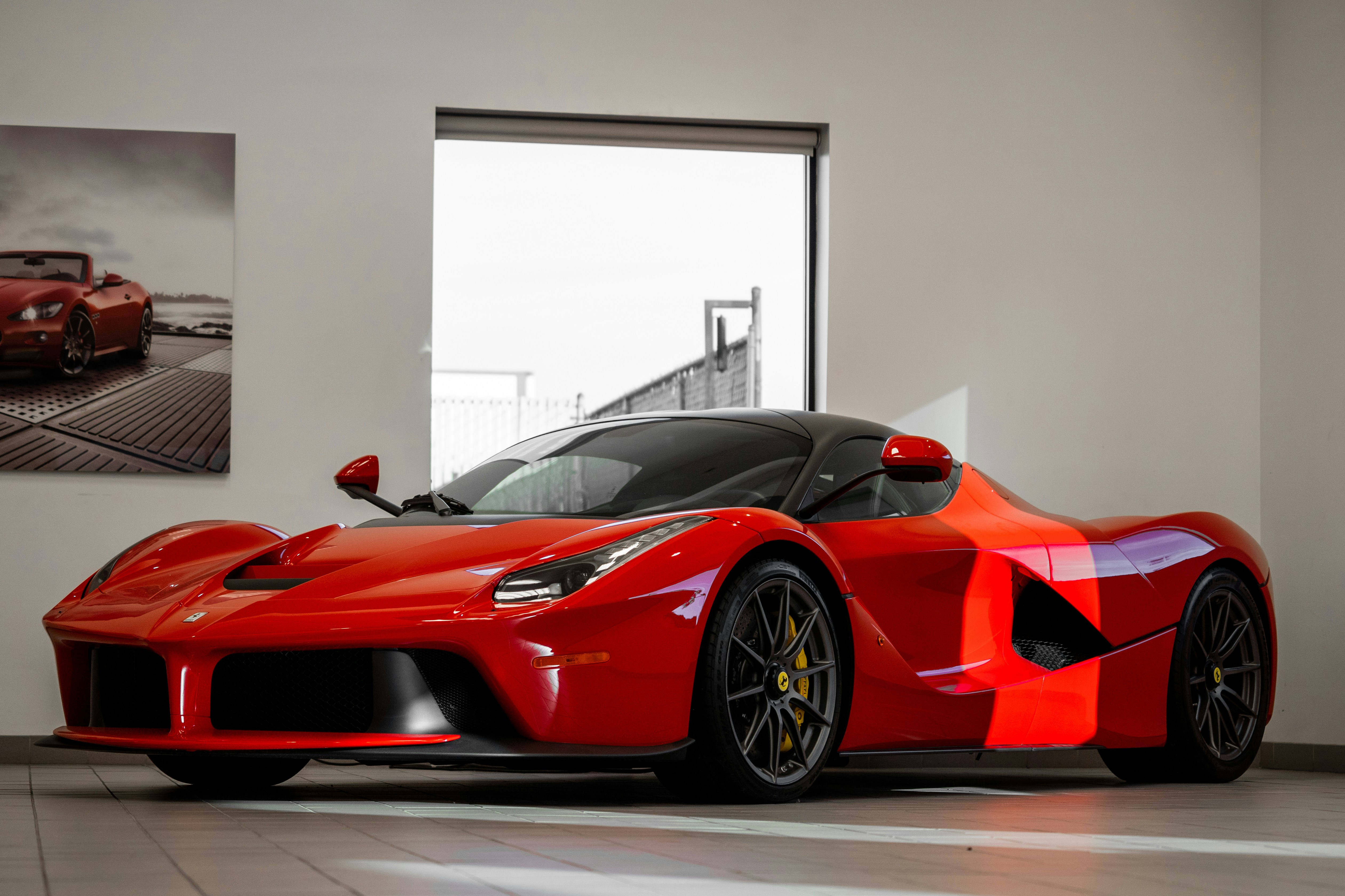Bright red Ferrari supercar displayed indoors in a showroom, featuring a low-slung aerodynamic body, black roof, sculpted side intakes, and large performance wheels under natural light.