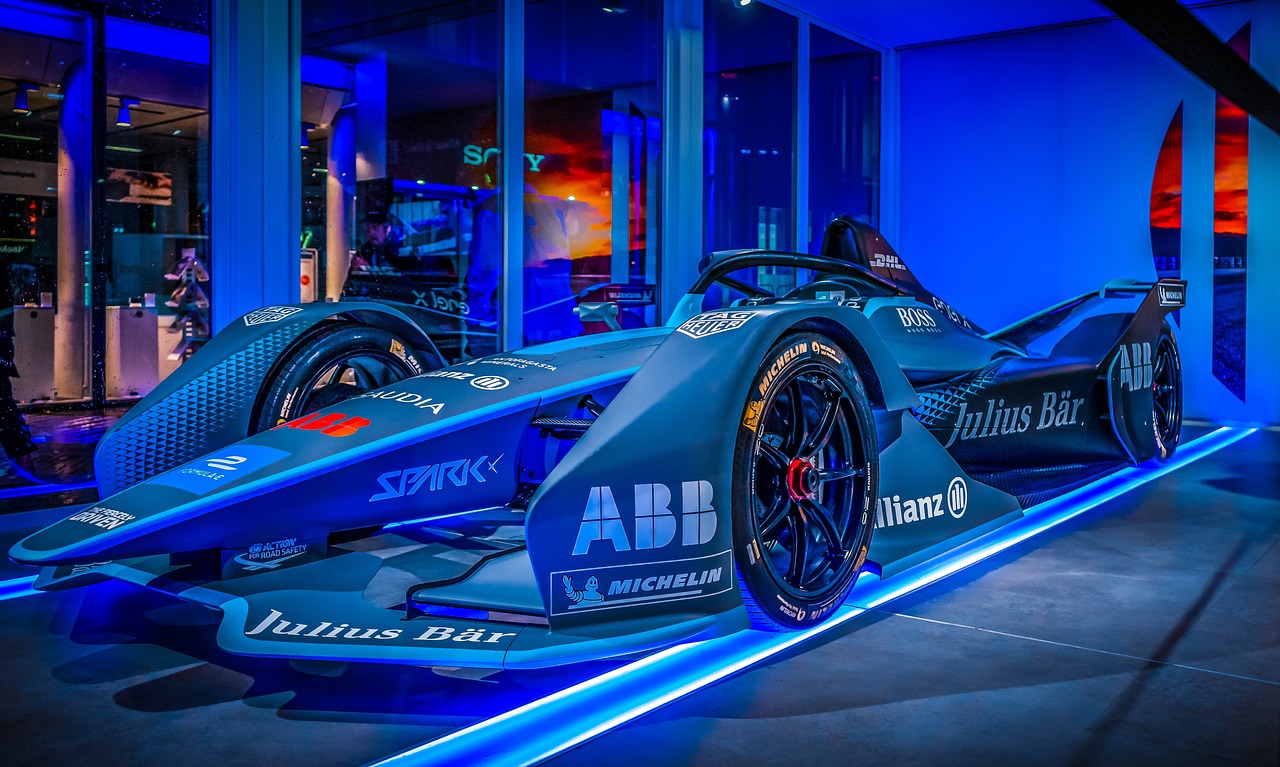 Electric Formula-style race car displayed indoors under blue accent lighting, featuring aerodynamic bodywork, exposed wheels, sponsor branding, and a futuristic exhibition setting.