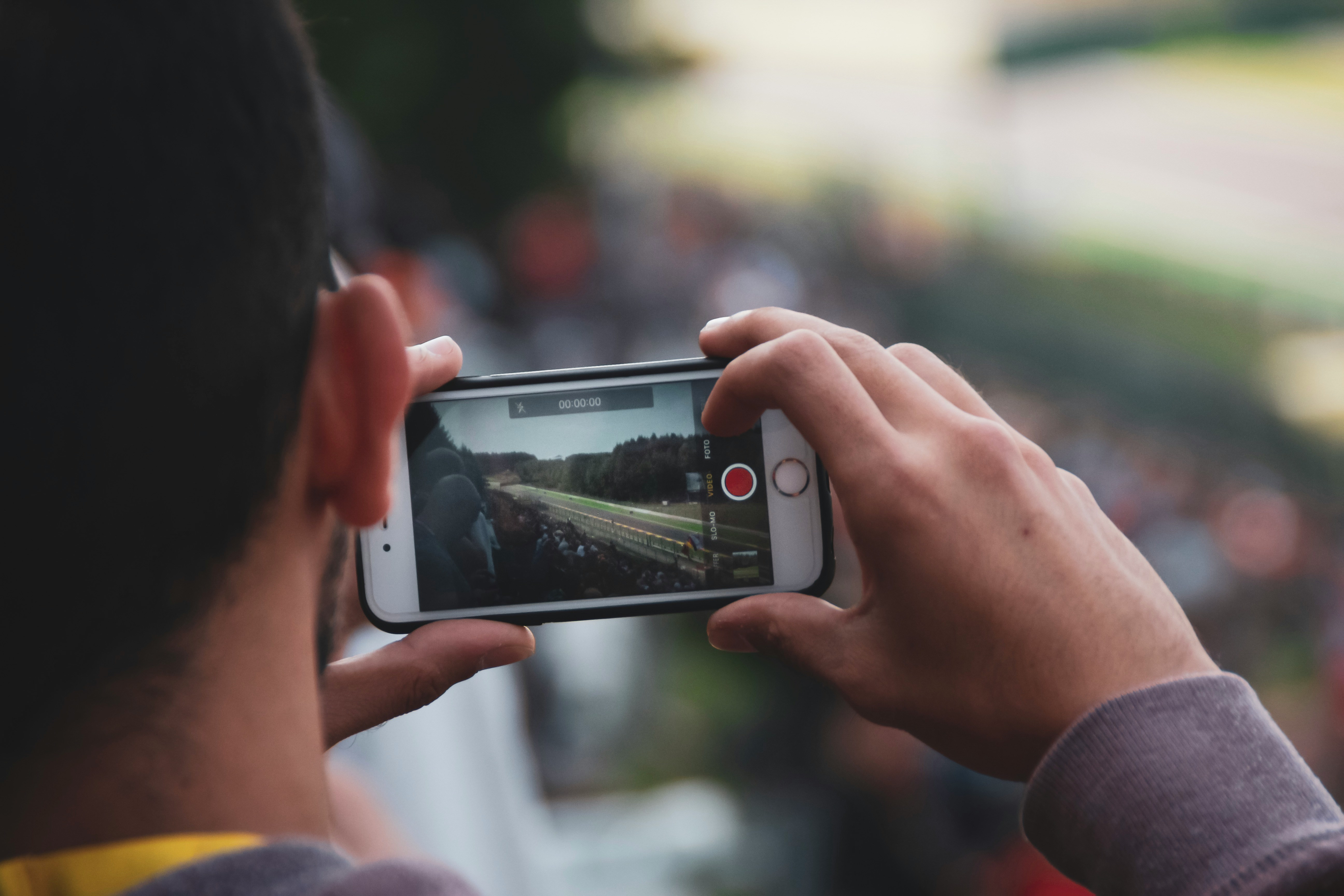 Person holding a smartphone horizontally to record or photograph an event, with the phone screen showing a crowd and activity in the distance while the background remains out of focus.