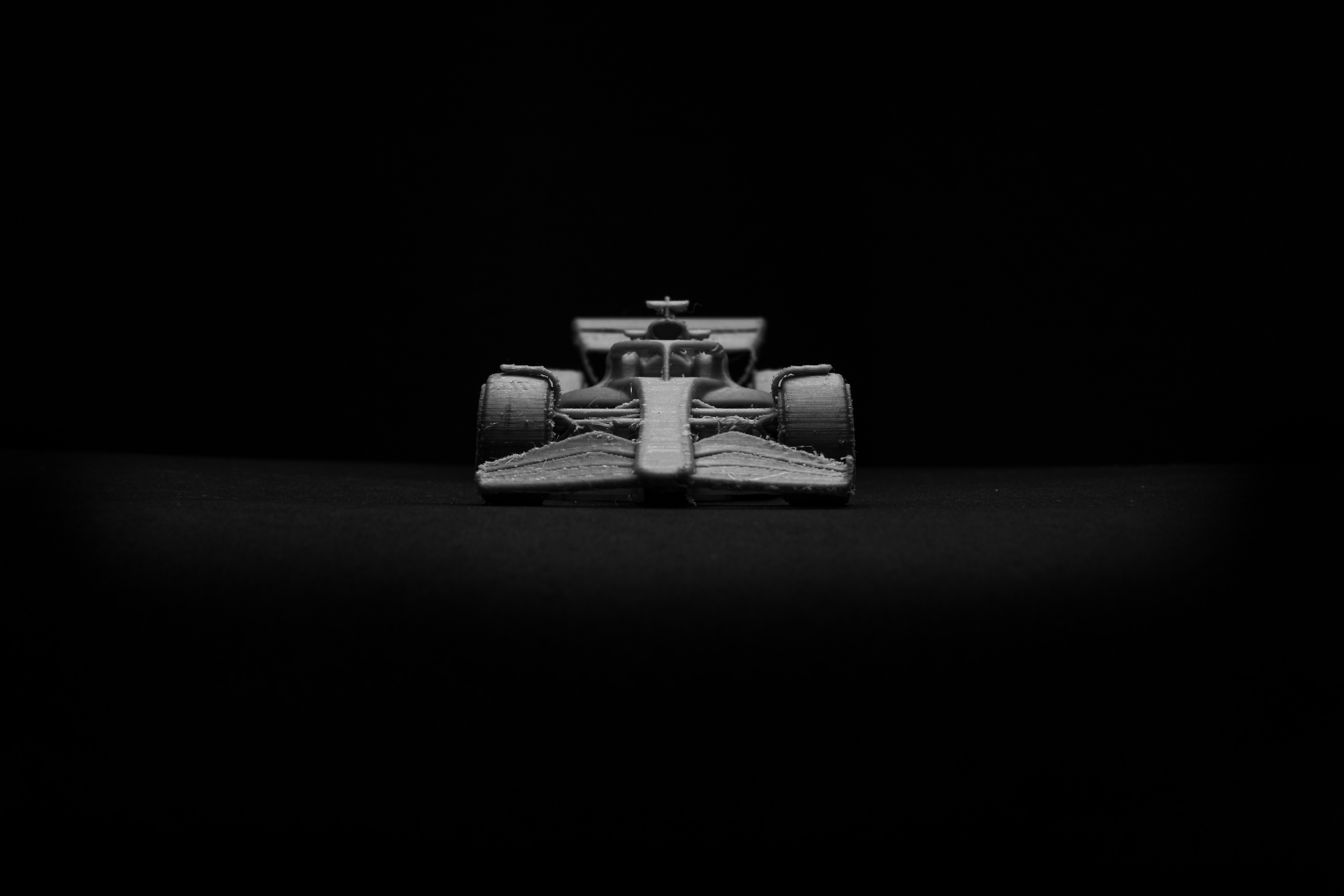 Front view of a Formula-style racing car positioned under dramatic studio lighting against a black background, with aerodynamic bodywork and wide racing tyres prominently displayed.