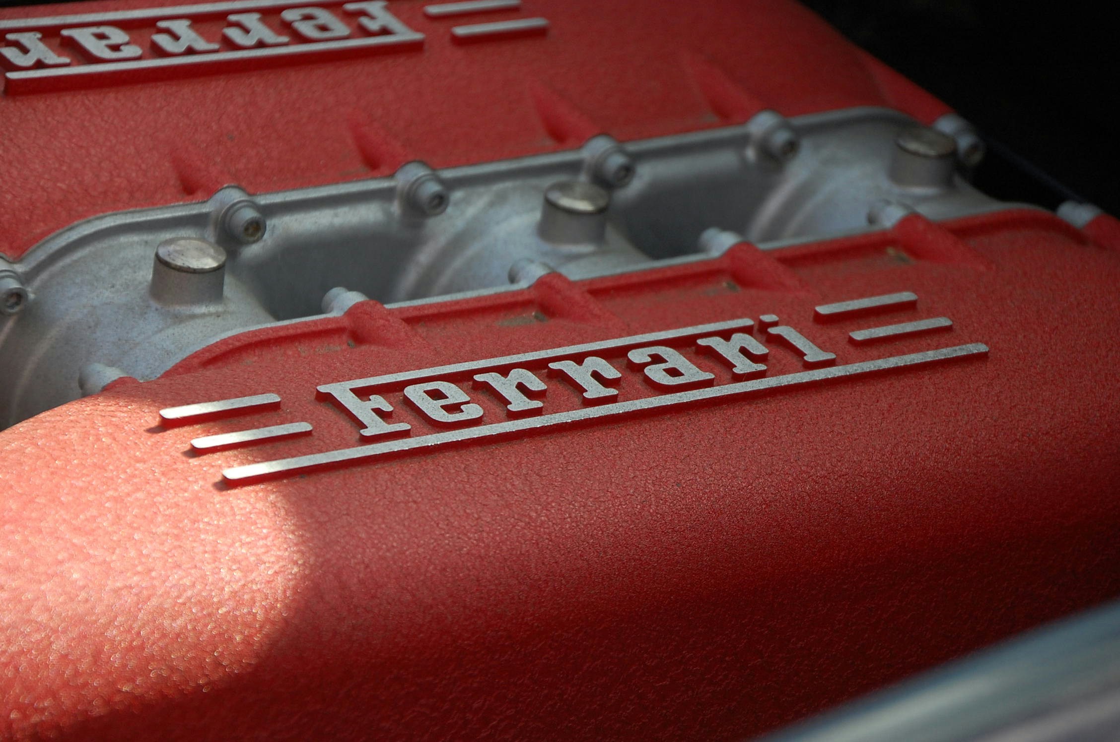 Close-up of a red Ferrari engine component featuring the raised “Ferrari” logo, with metallic engine parts visible in the background.